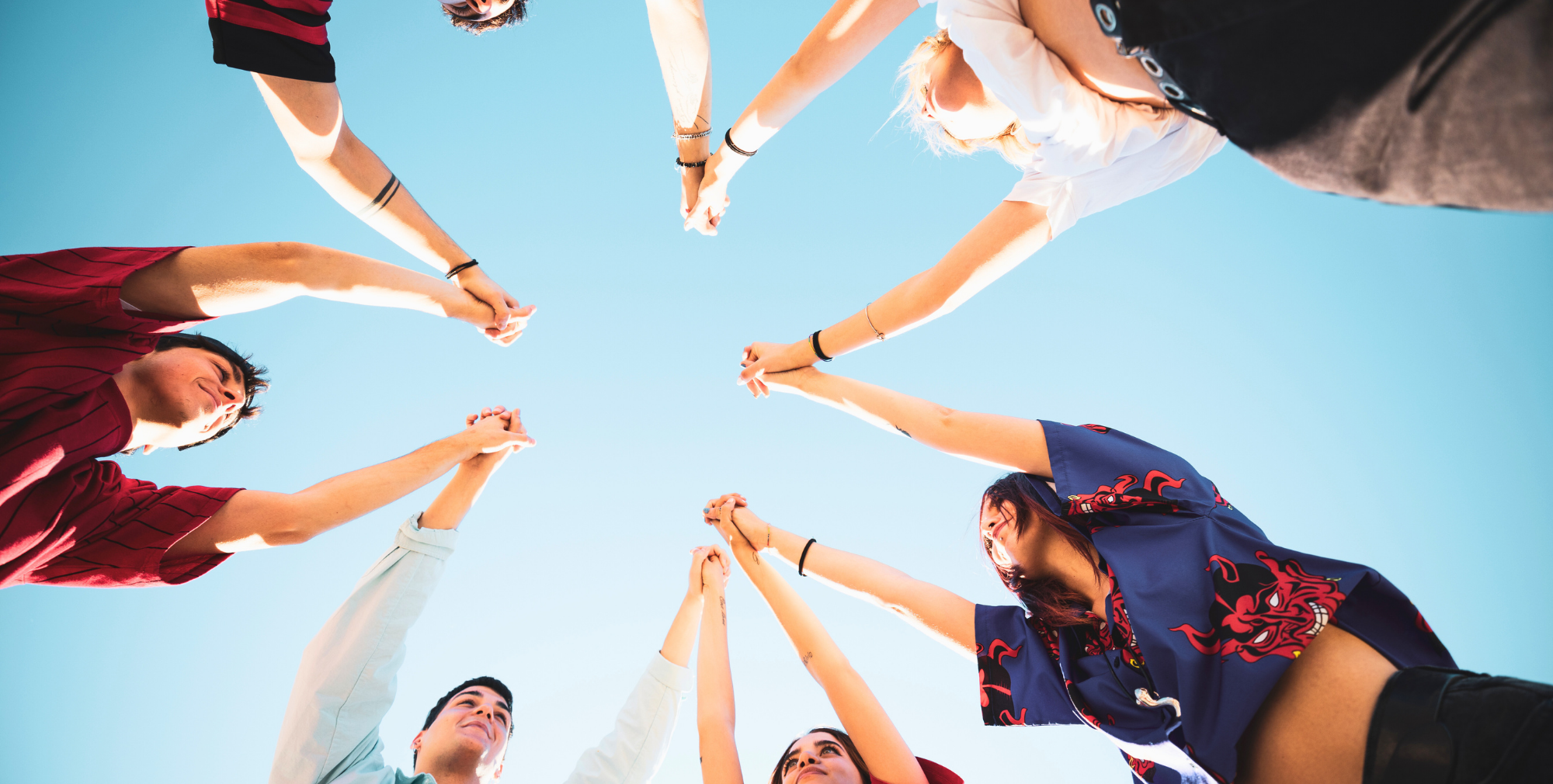 Group of young people standing in a circle, holding hands from a low-angle view against a blue sky.