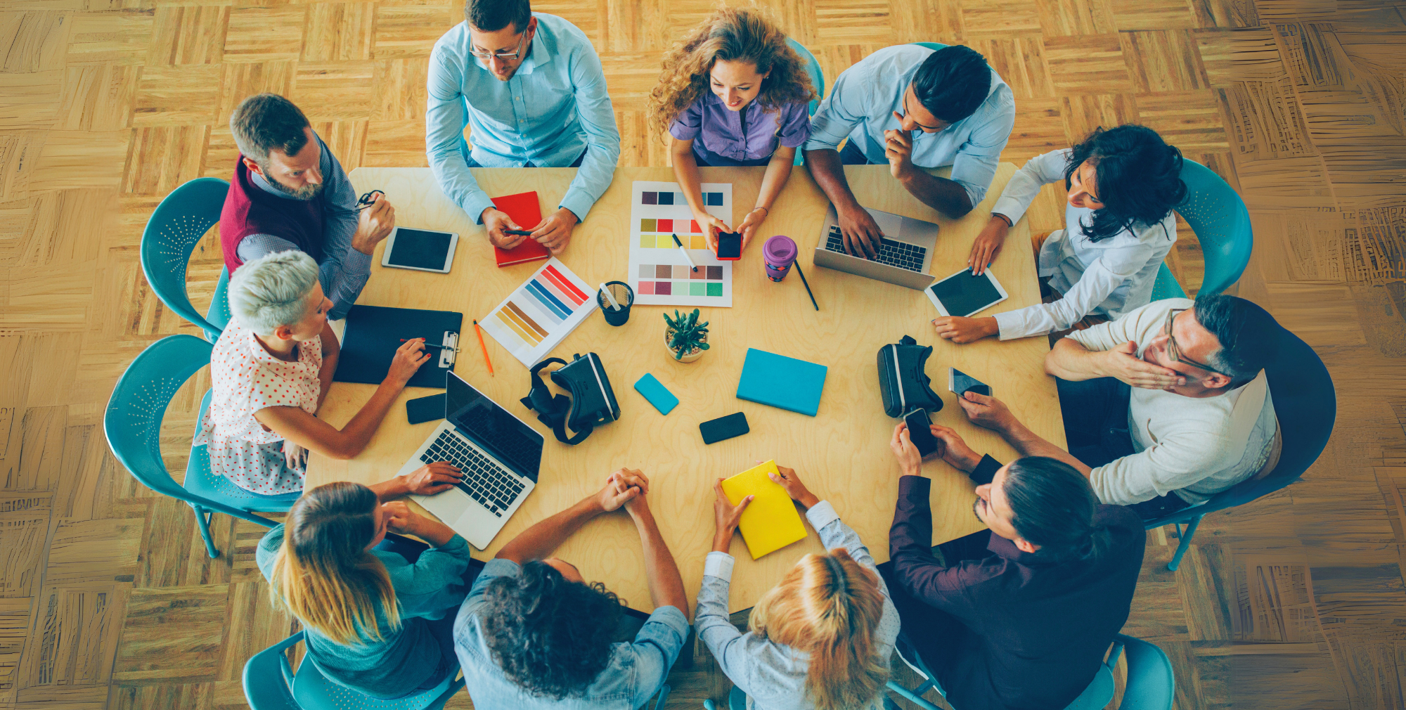A group of professionals gathered around a large table, collaborating on a creative project with laptops, colour swatches, notebooks, and tablets.