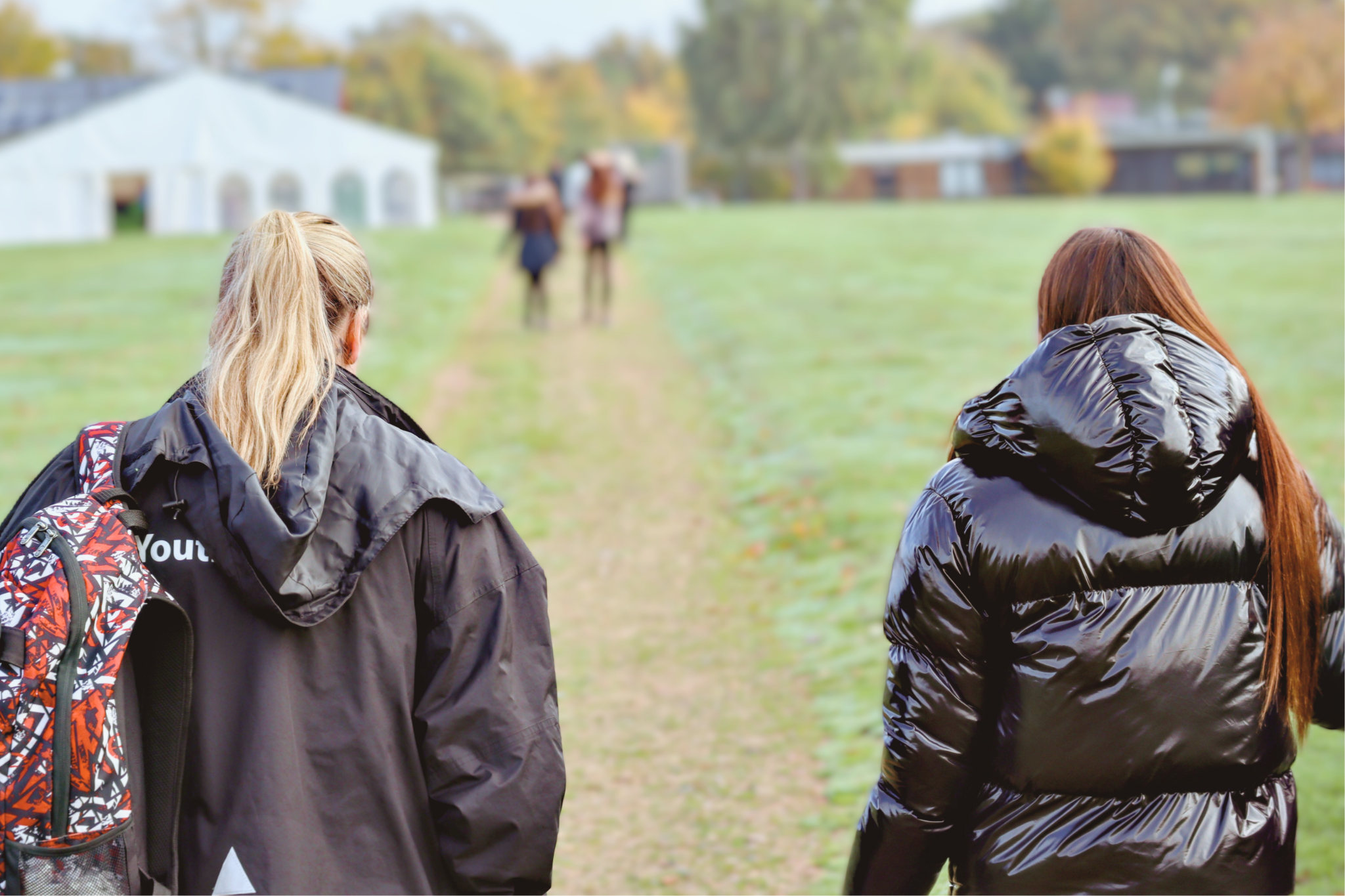 Two people walking on a grassy path toward a large white marquee in the distance, with autumn trees and buildings in the background. One carries a colourful backpack.
