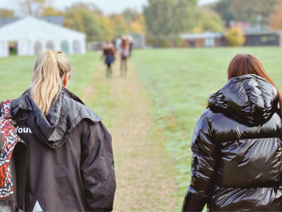 Two people walking on a grassy path toward a large white marquee in the distance, with autumn trees and buildings in the background. One carries a colourful backpack.