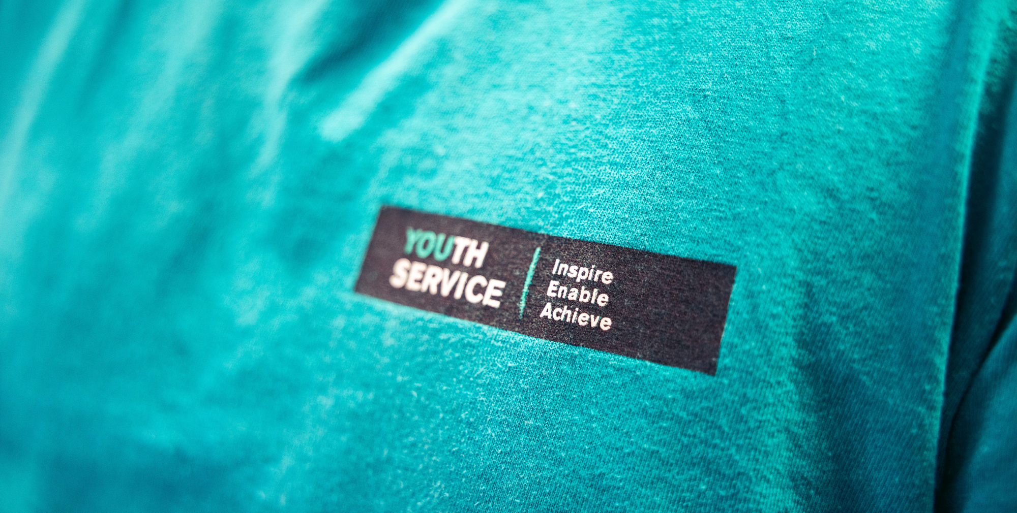 Youth Service logo on a teal staff shirt displaying the words Inspire, Enable, Achieve