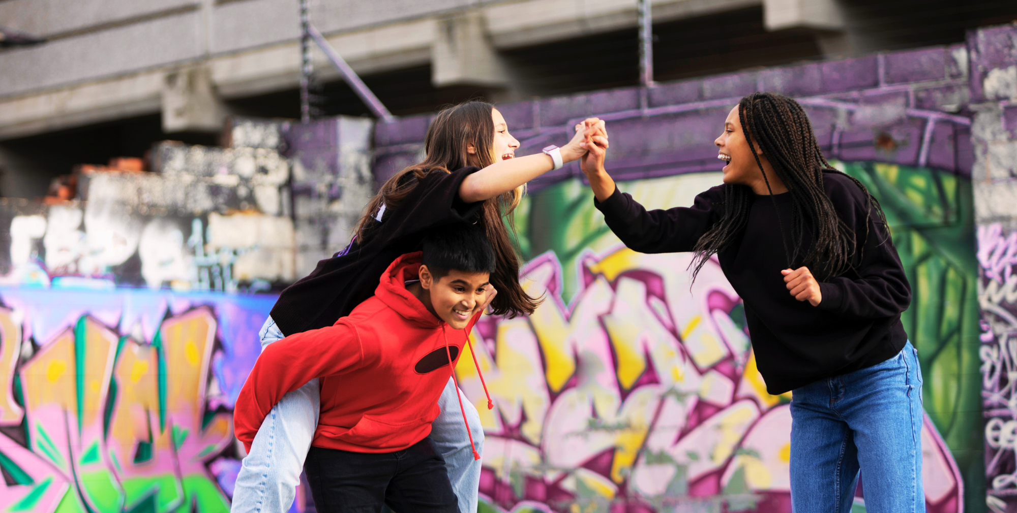 Three young people smiling and playing in front of colourful graffiti — one giving a piggyback while the other two high-five with laughter