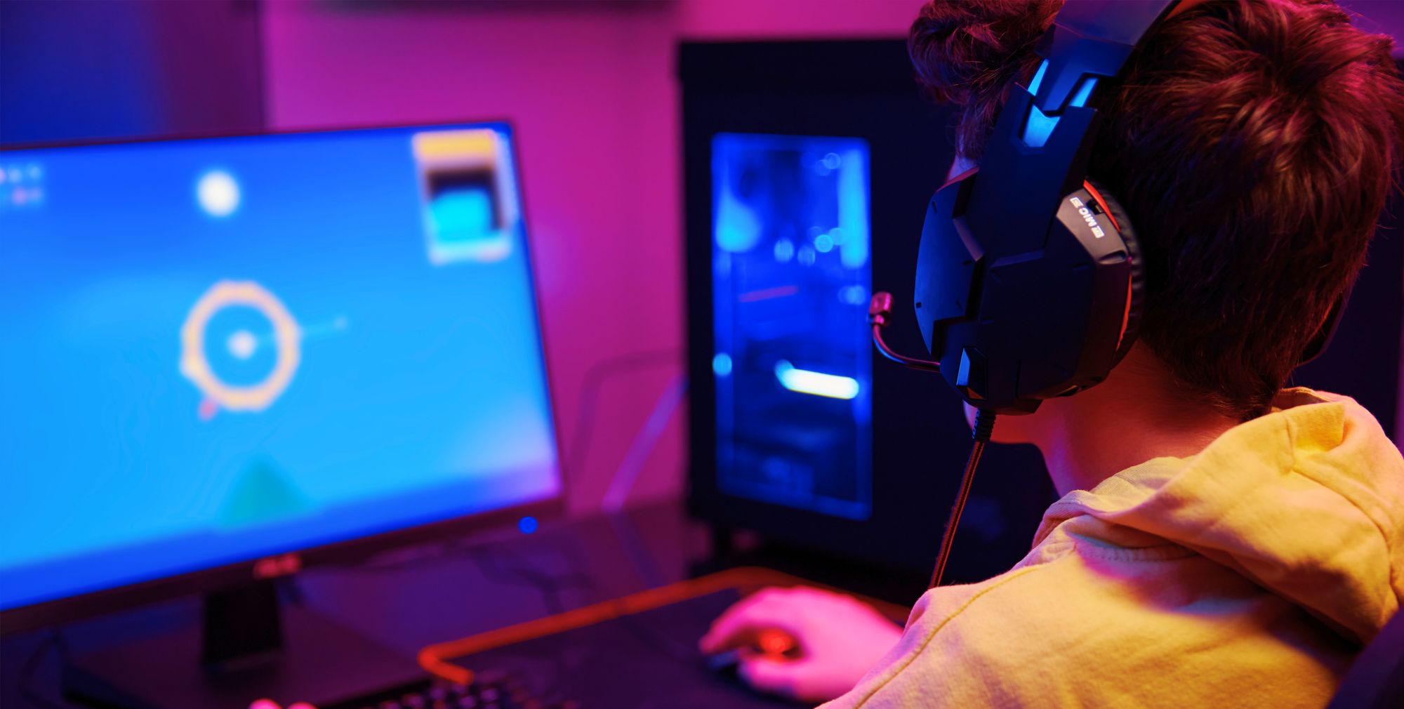 A young person wearing headphones, focused on a computer screen with a colourful game interface in a dimly lit, neon-coloured room