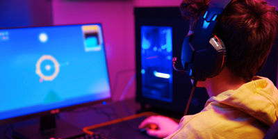 A young person wearing headphones, focused on a computer screen with a colourful game interface in a dimly lit, neon-coloured room