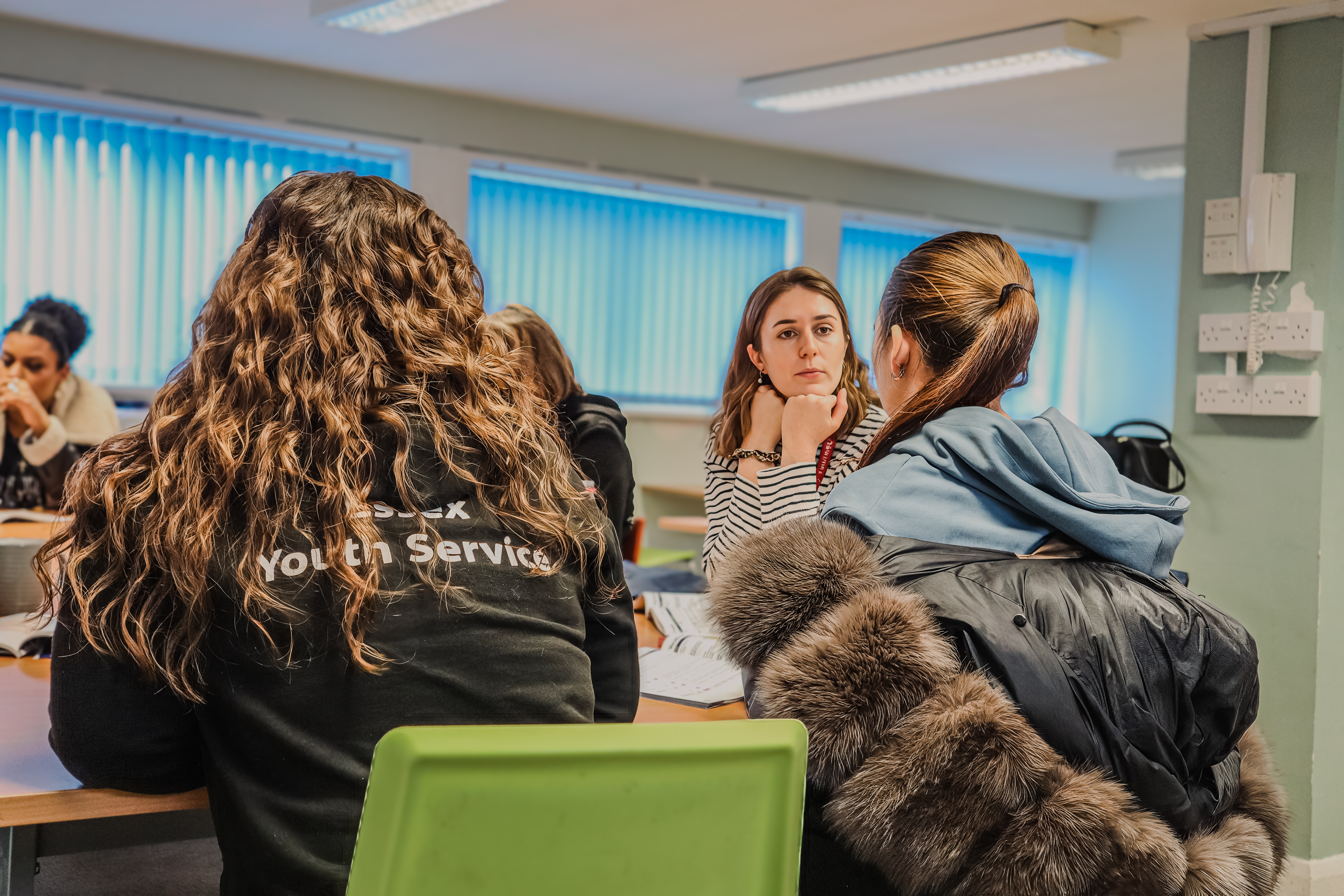 A young women sitting at a table in conversation with a female youth worker, who is attentively listening. Another woman is wearing an ‘Essex Youth Service’ jacket.